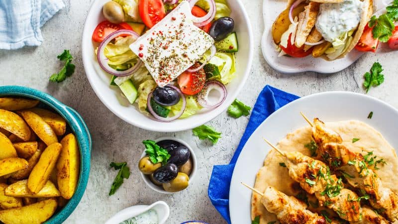 A spread of Greek food including salad, olives, potato wedges, and chicken souvlaki.