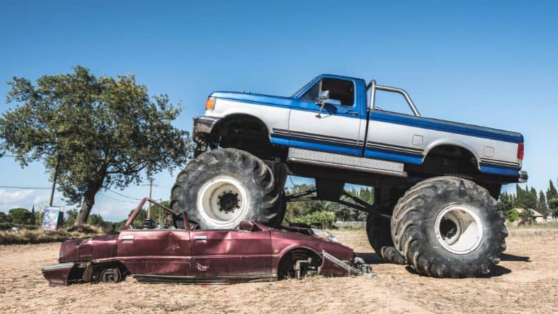 A monster truck crushes a smashed red car in an open dirt field.