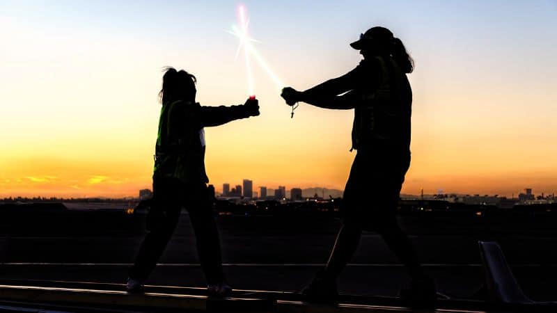 Two people in safety vests playfully dueling with toy lightsabers at sunset.