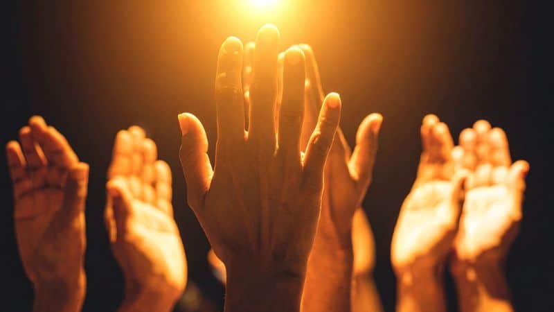 Several hands reach upward toward a warm, glowing light.