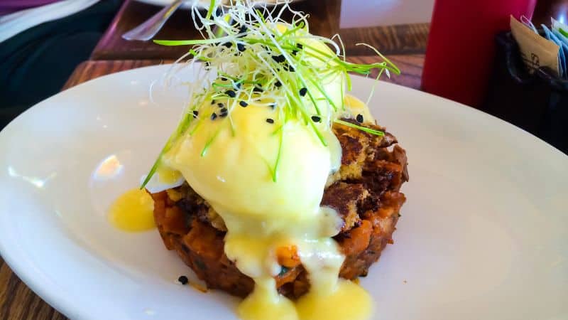 Close-up of a gourmet brunch dish featuring a crab cake topped with a poached egg and drizzled with hollandaise sauce, garnished with sprouts and black sesame seeds on a white plate. The dish is presented in a casual dining setting with hints of other table items blurred in the background.