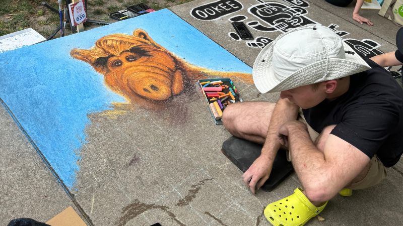 An artist at a sidewalk arts festival creating a vibrant chalk drawing of ALF, the TV show character, on the pavement. The artist, wearing a light-colored bucket hat and yellow crocs, is crouched beside his artwork, looking thoughtfully at his selection of chalks laid out next to him. The artwork is prominently displayed with a clear blue background.