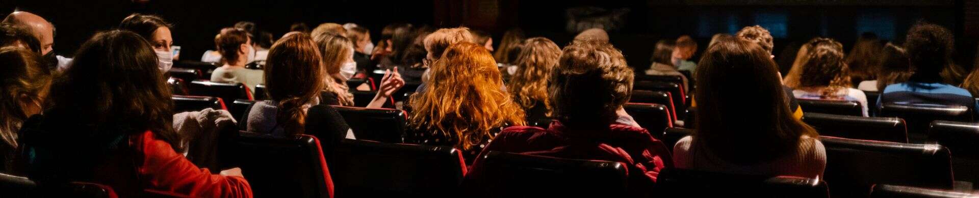 A crowded theater audience viewed from behind, with people sitting in rows of red and black chairs.