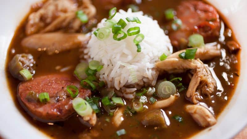A bowl of gumbo with sausage, shredded chicken, and a mound of white rice topped with green onions.