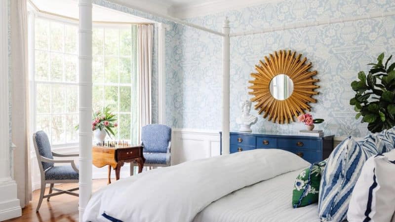 Bright bedroom with a white four-poster bed, blue floral wallpaper, and a sunburst mirror above a blue dresser.