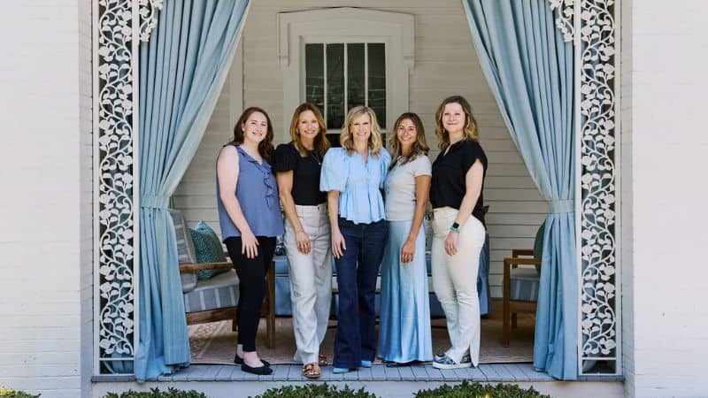 Five women standing on a porch framed by light blue curtains and decorative trim.