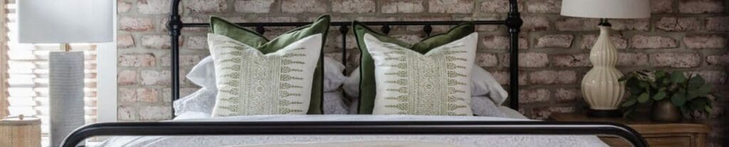 A cozy bedroom with a black metal bed frame, green and white accent pillows, and brick wall backdrop.