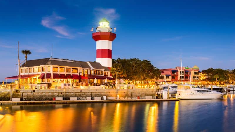 Red and white striped lighthouse overlooking a marina with boats at dusk.