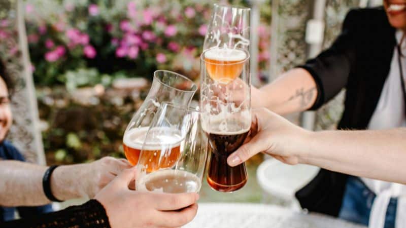 Friends clinking glasses of beer at an outdoor table with flowers in the background.