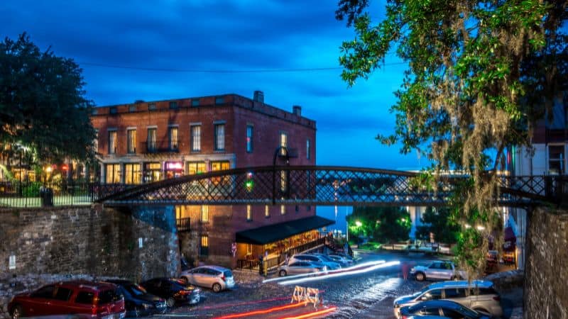 An iron pedestrian bridge over a cobblestone street at dusk, with a historic brick building and Spanish moss-draped trees in Savannah's River Street district.