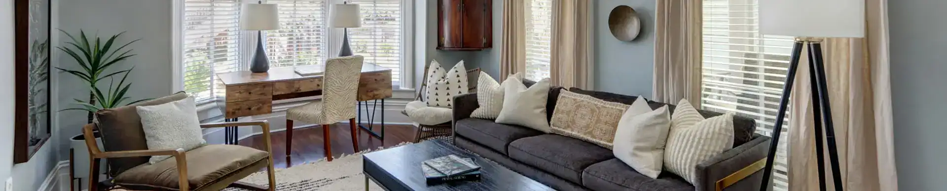 The Taylor House living room with a gray sofa, a wooden desk, and tall windows with soft curtains