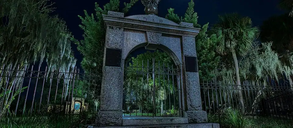Historic stone cemetery gate lit up at night, framed by Spanish moss and oak trees