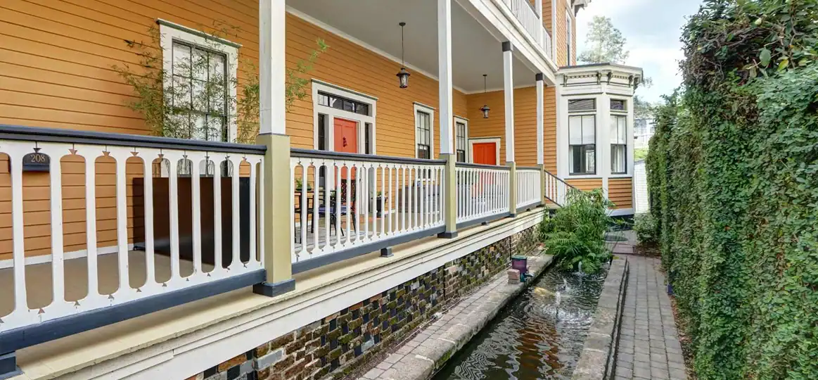 Orange clapboard house with a white-railed porch and red doors, beside a brick walkway with a narrow water channel and a tall green hedge