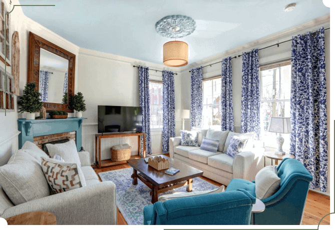 Living room with white sofas, blue floral curtains, a teal fireplace mantel, a large wood-framed mirror, and a woven pendant light